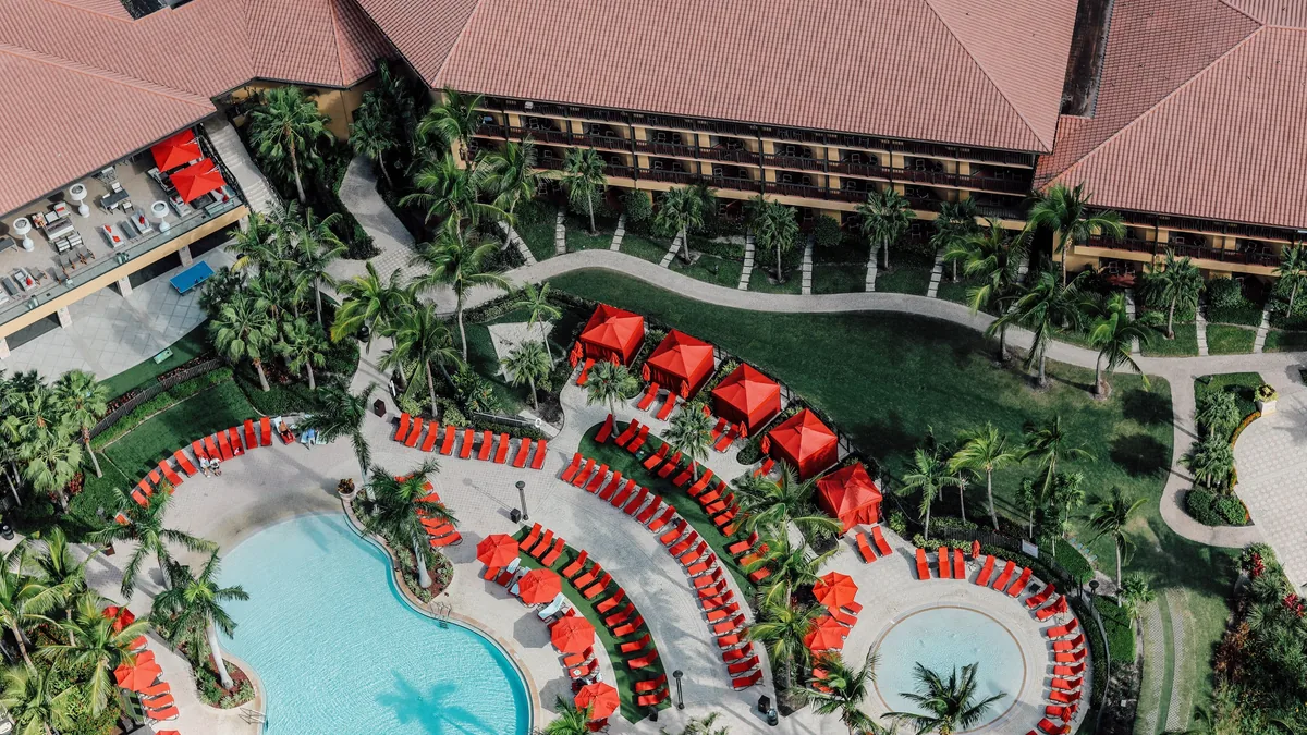 This aerial photograph depicts a resort with a large pool, surrounded by palm trees and red lounge chairs, featuring a building with a brown